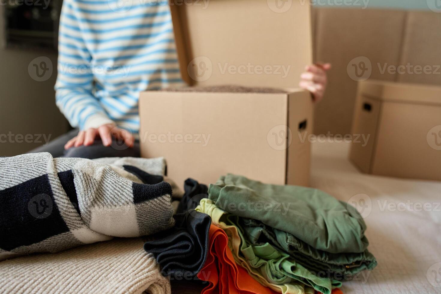 Female packs donation box with clothes at home. Person places warm sweater into cardboard box. Preparing used clothing for recycling or second hand. Concept of charity and humanitarian aid photo