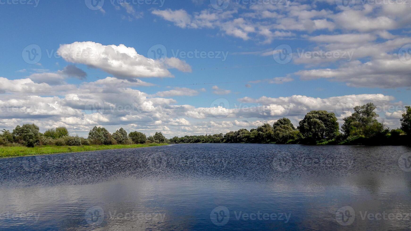 Serene summer landscape with a calm river, lush green trees on both sides and a blue sky filled with soft white clouds. Tranquil natural scene with clear reflections in the water. photo