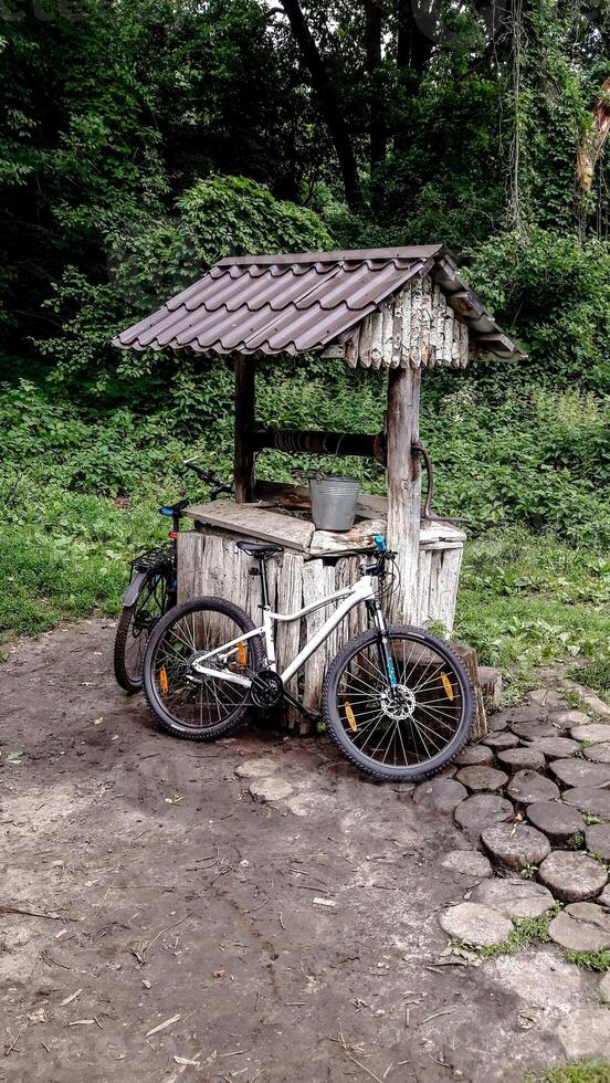Two bicycles leaning against an old wooden well in the middle of a green forest. Peaceful summer scene with natural textures, trees, and tranquil atmosphere. photo