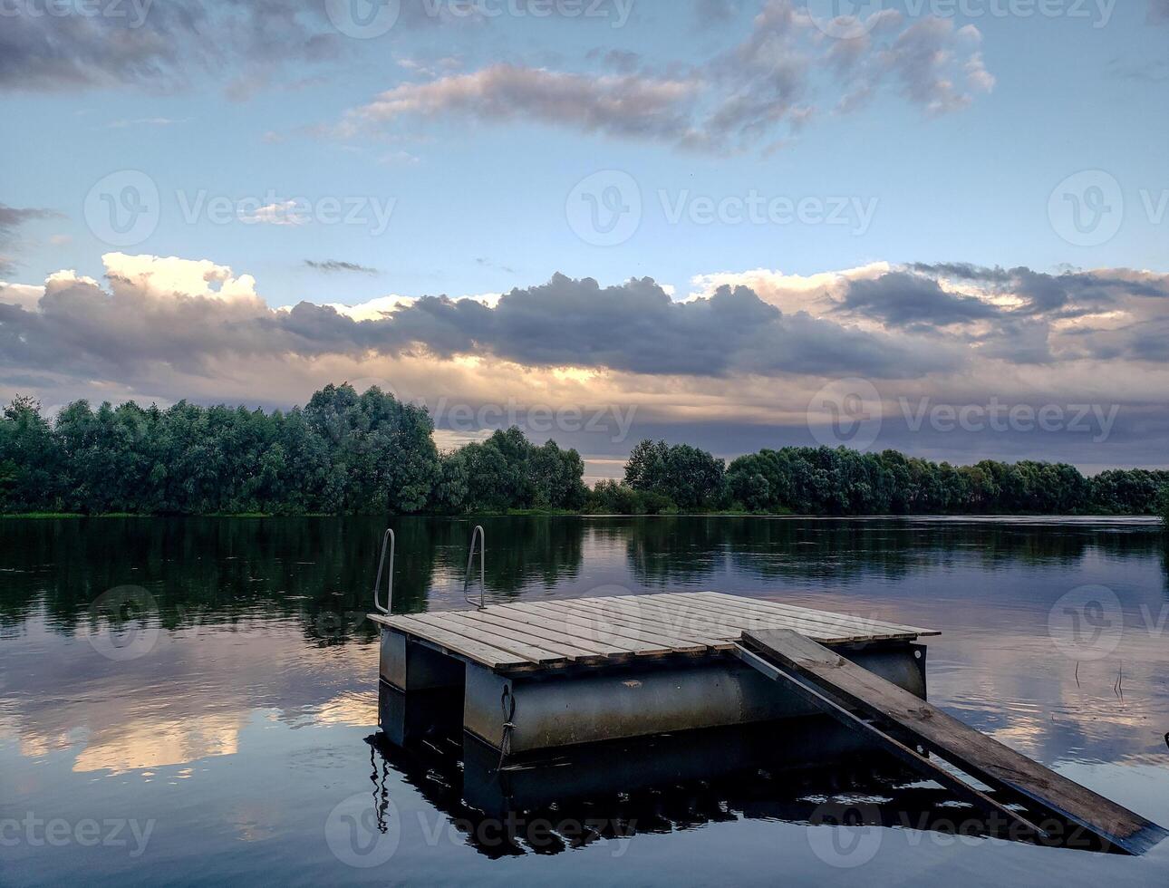 Peaceful rural river scene with wooden raft, sunset sky and forest reflected in calm water. Natural landscape with soft colors and atmospheric clouds. photo