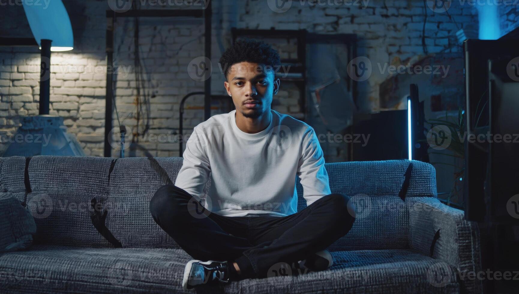 Young man sits cross-legged on a couch in a dimly lit room with blue lighting and an industrial background photo