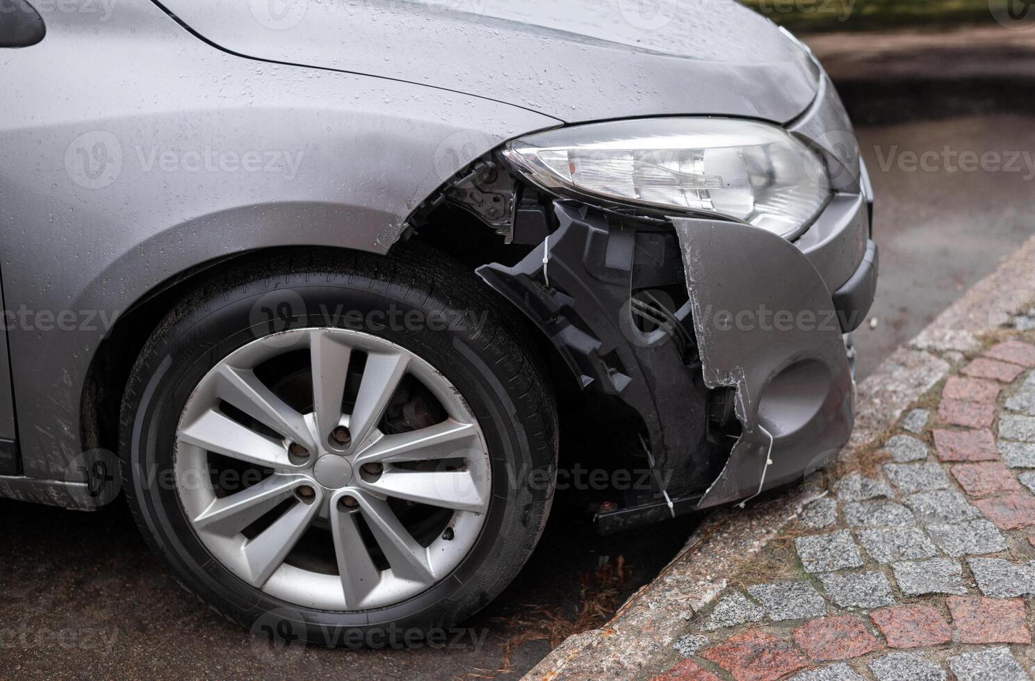 Front of a silver car with a damaged bumper and exposed inner structure after an accident, parked near a curb on a wet road photo