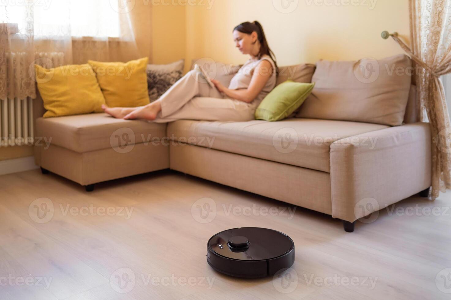 A robot vacuum cleaner washes the floor while a young woman reads a book sitting on the sofa. Smart home, automation and modern technologies in everyday life photo