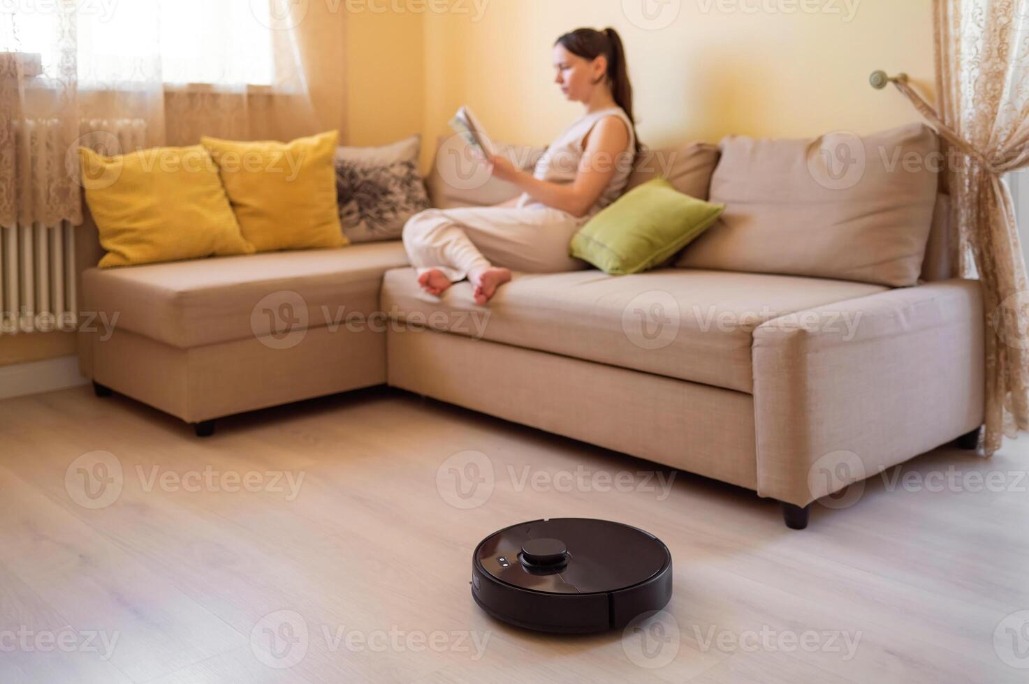 A robot vacuum cleaner washes the floor while a young woman reads a book sitting on the sofa. Smart home, automation and modern technologies photo
