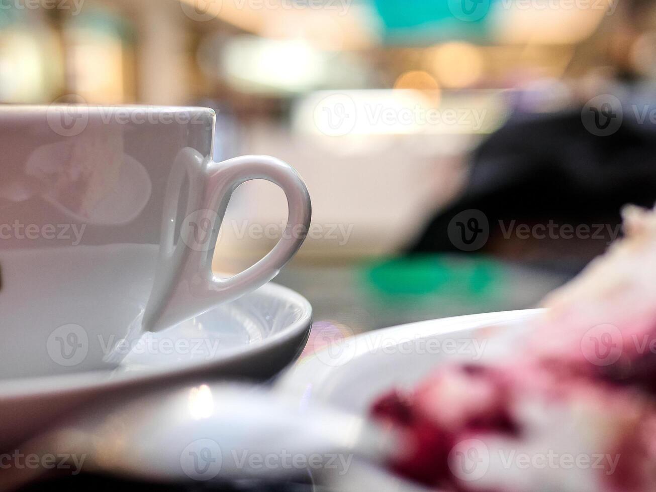 A white cup with a handle sits on a saucer next to a plate of cake photo