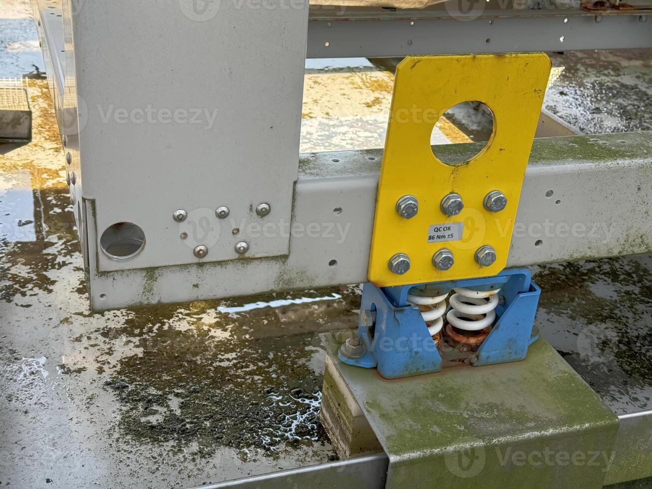 A close-up view of a mechanical spring assembly mounted on a metal frame, showing a yellow and blue color scheme. The area is slightly dirty with water stains on the surface. photo