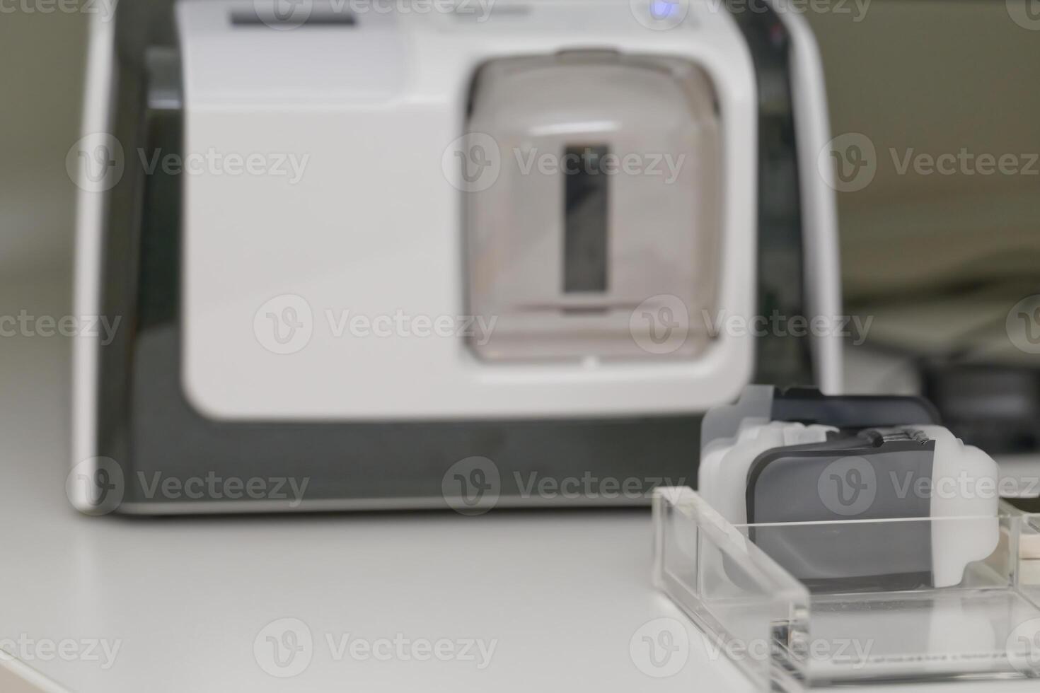 A closeup view of a medical device xray scanner with a transparent chamber and various attachments. The background features cables and other equipment, suggesting a clinical setting. photo