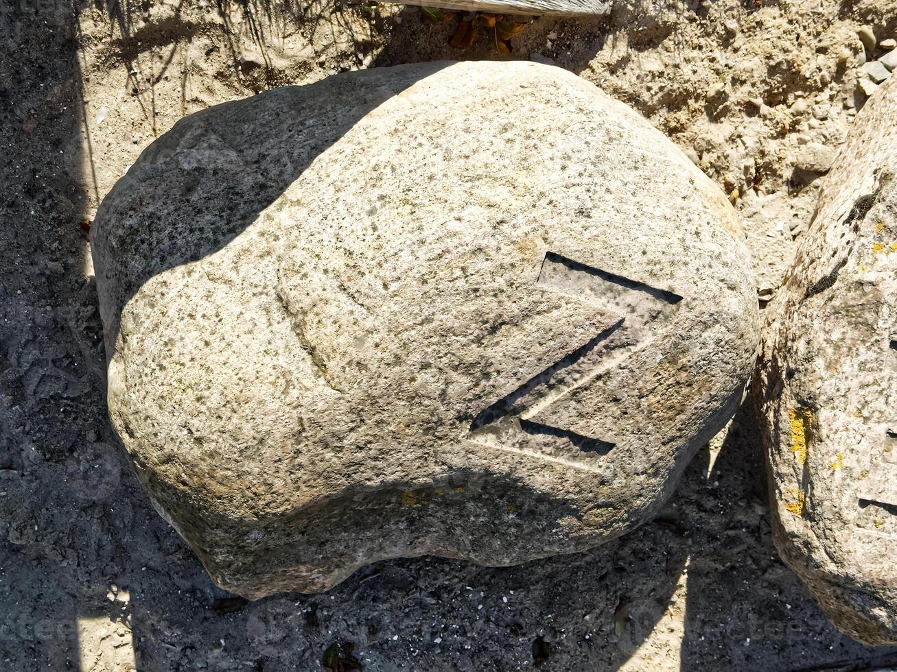 A large, weathered rock with a distinct carved symbol resembling the letter 'Z' on its surface, surrounded by sandy ground. photo