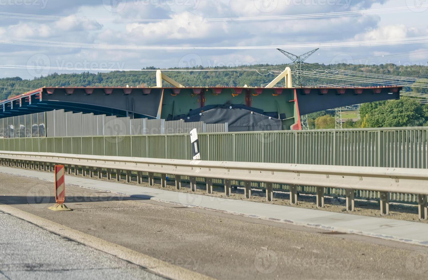 A view of a large, modern building with a distinctive roof design, partially obscured by a fence. The scene is set alongside a highway, with construction barriers visible in the foreground. photo