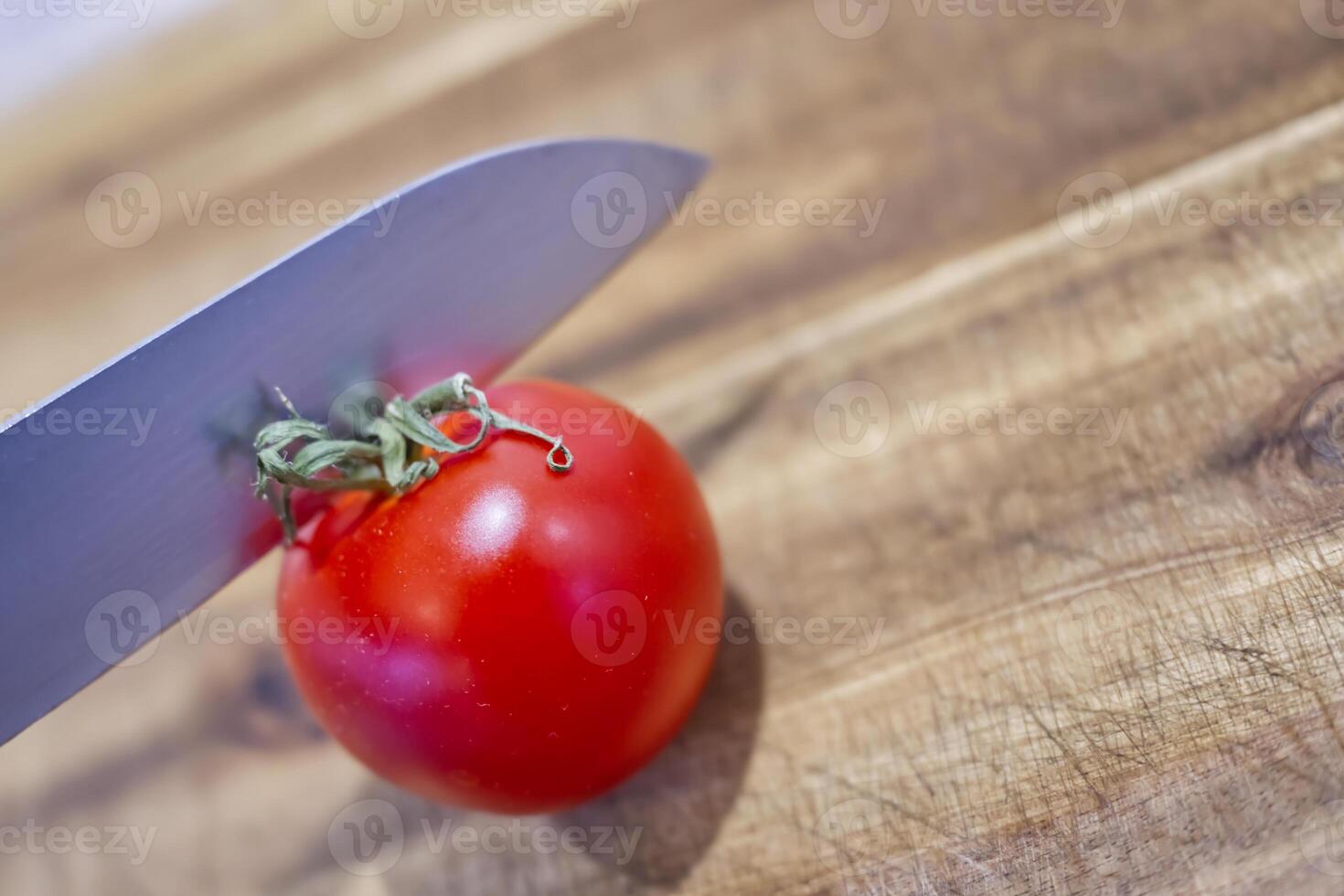 A close-up of a shiny red tomato on a wooden cutting board, with a sharp knife poised above it, ready to slice. photo