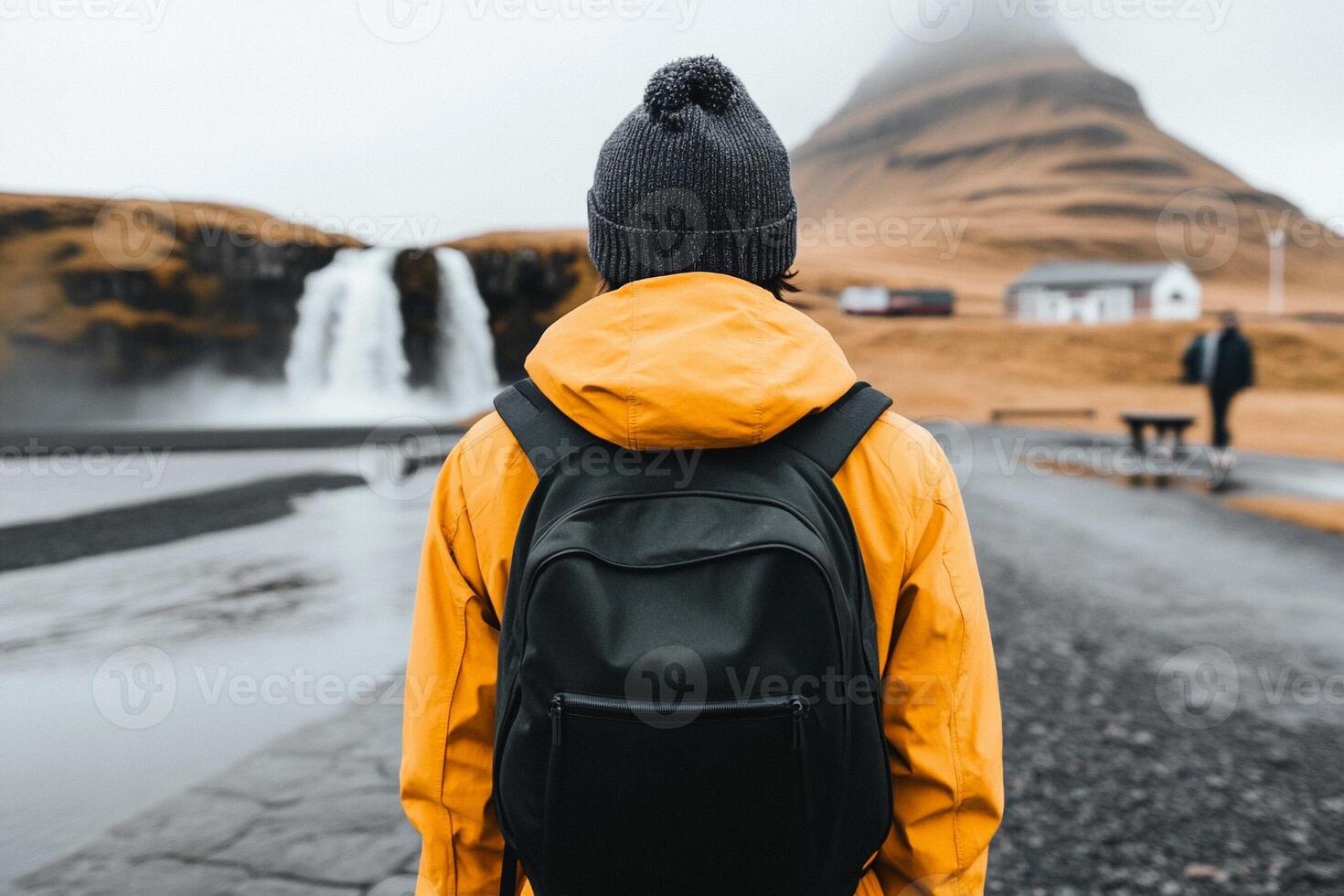 A man in a yellow jacket and black backpack standing in front of a waterfall photo