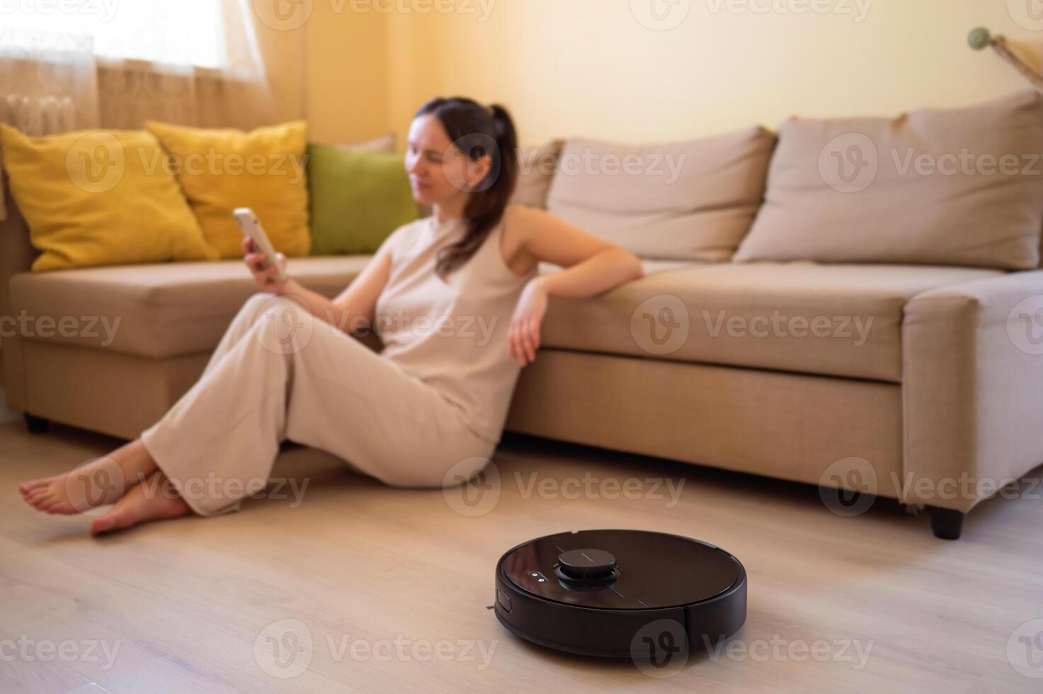 A robot vacuum cleaner washes the floor while a young woman uses her smartphone sitting on the floor next to the sofa. Smart home, automation and modern technologies in everyday life photo