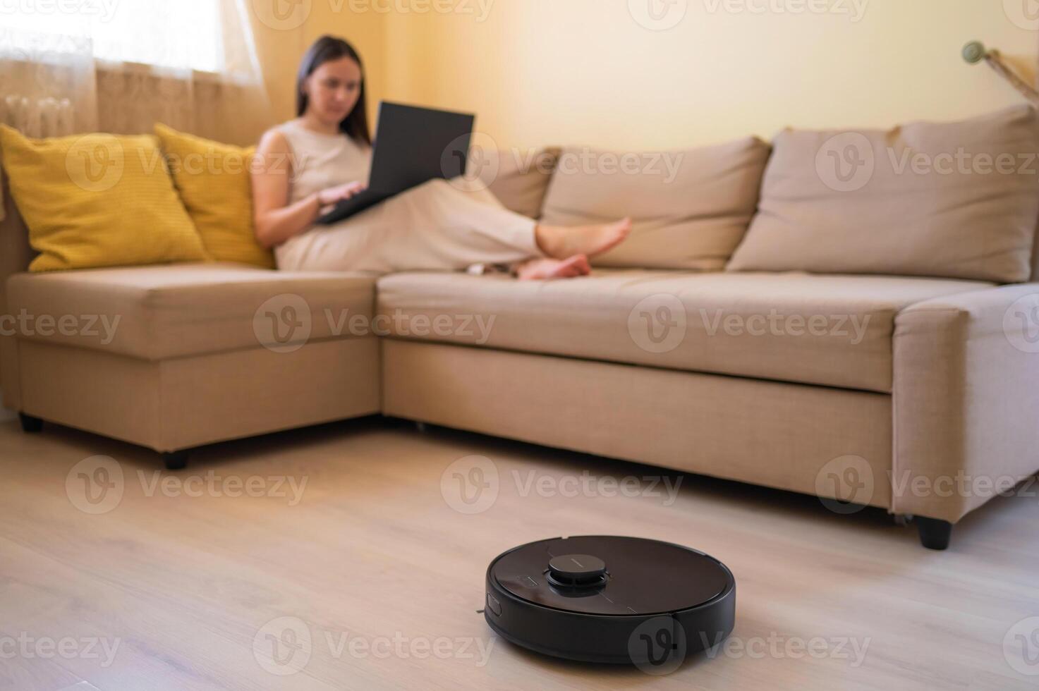 A robotic vacuum cleaner cleans the floor while a young woman works on her laptop, sitting on a cozy beige sofa. Smart home, automation, and modern technology in daily l photo