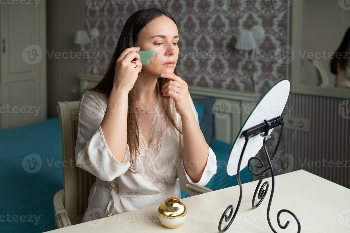 A young woman in a silk robe uses a jade gua sha facial tool photo
