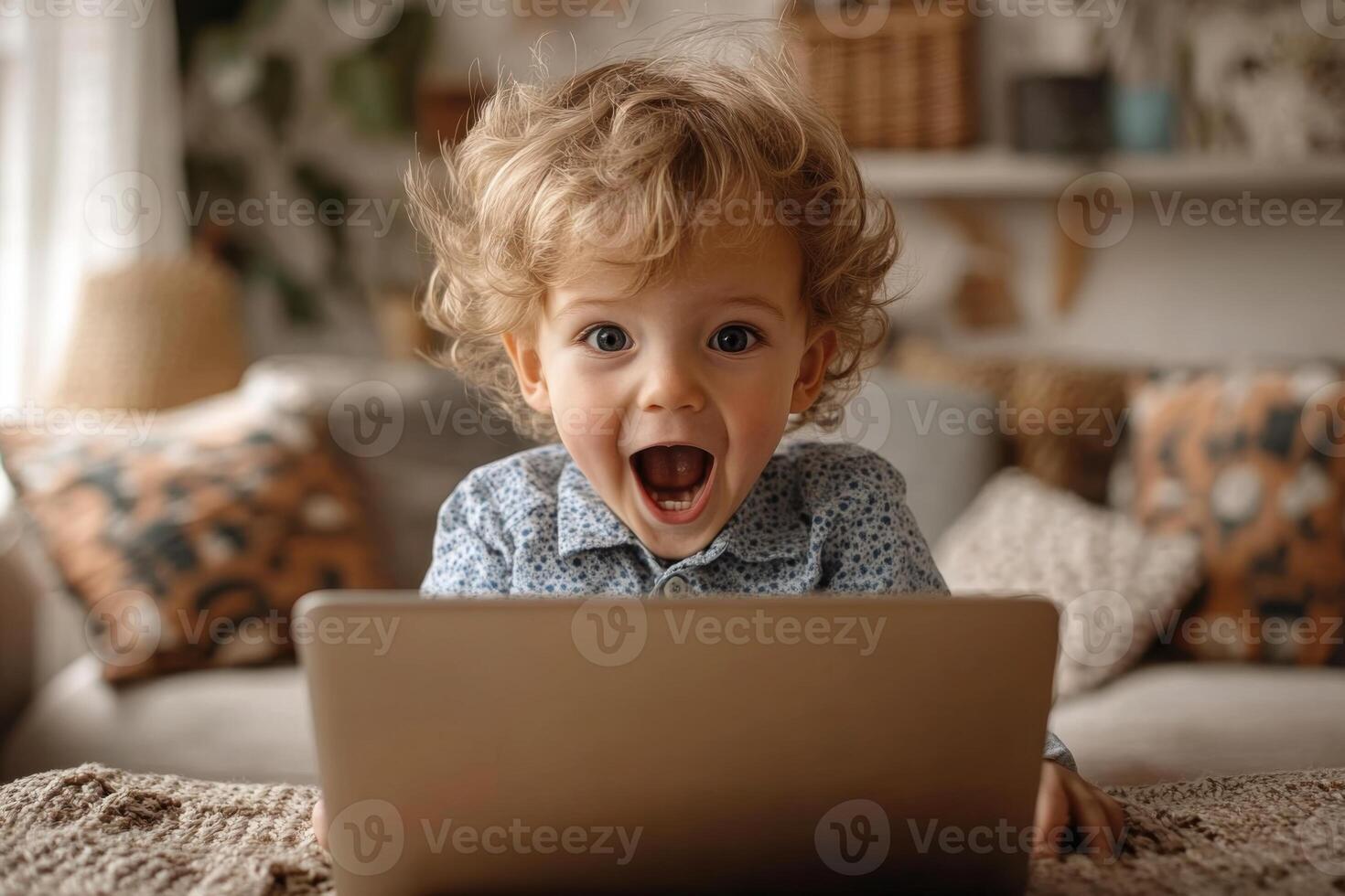 Small child looks surprised at the laptop screen. He is at home in a living room, sitting on a couch with pillows. photo