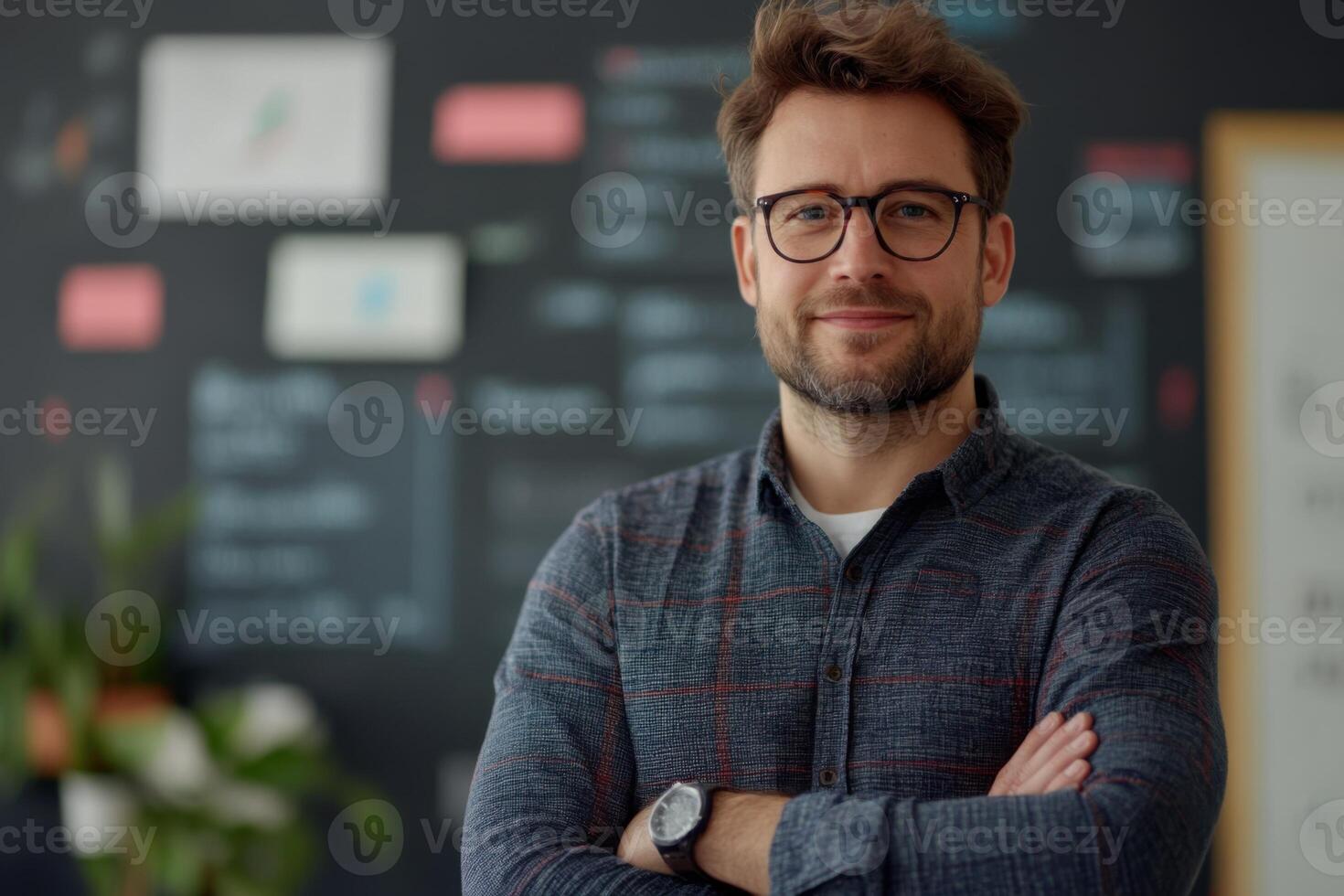 Person stands with arms crossed. He wears glasses. A blackboard with charts is behind. He has a watch. The lighting is bright. photo