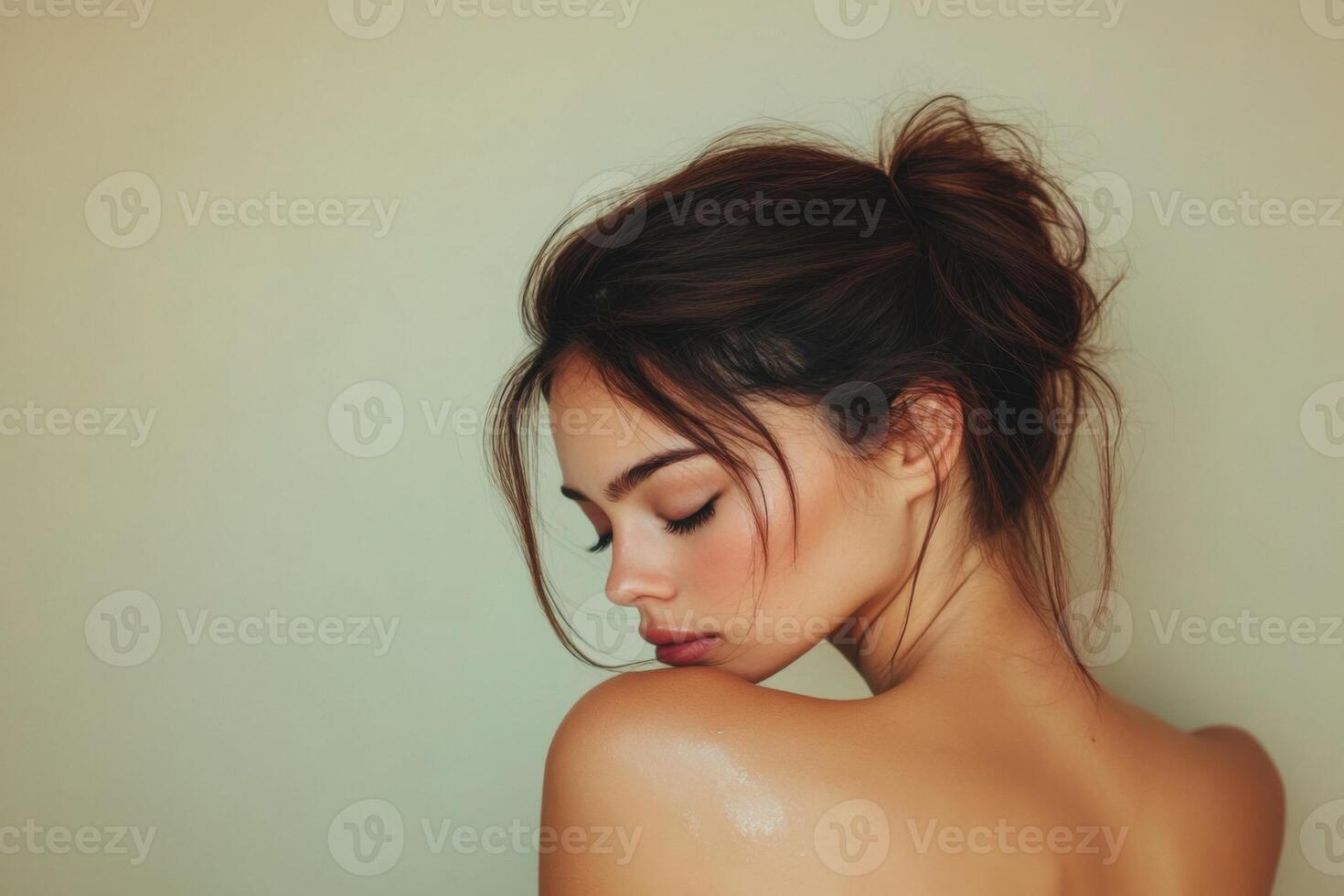 Profile of a beautiful woman with dark hair in a messy bun. Soft skin and gentle light against a plain neutral wall. photo