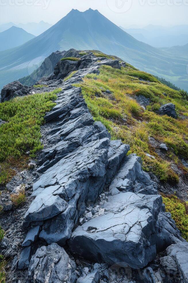 Rocky mountain ridge leading to distant peak. Green plants grow along the rocky path. Mountain range fades into the background. Outdoor scene. photo