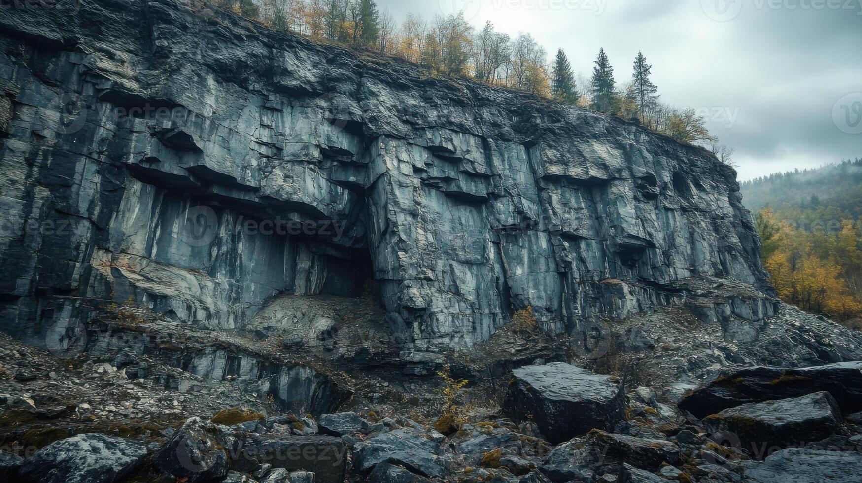 Steep stone wall with a cave opening and scattered rocks below. Cloudy sky above. Autumn foliage on trees atop the wall and nearby hillside. photo