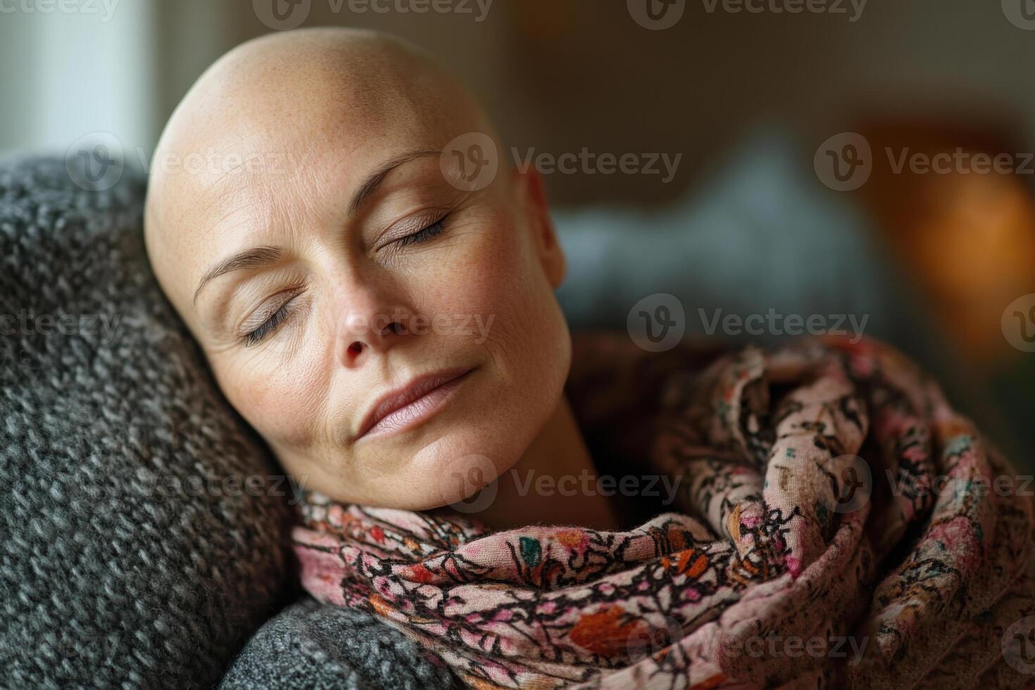 Woman with shaved head rests peacefully. She is sleeping and wearing a colorful scarf while leaning on a gray textured pillow indoors. photo