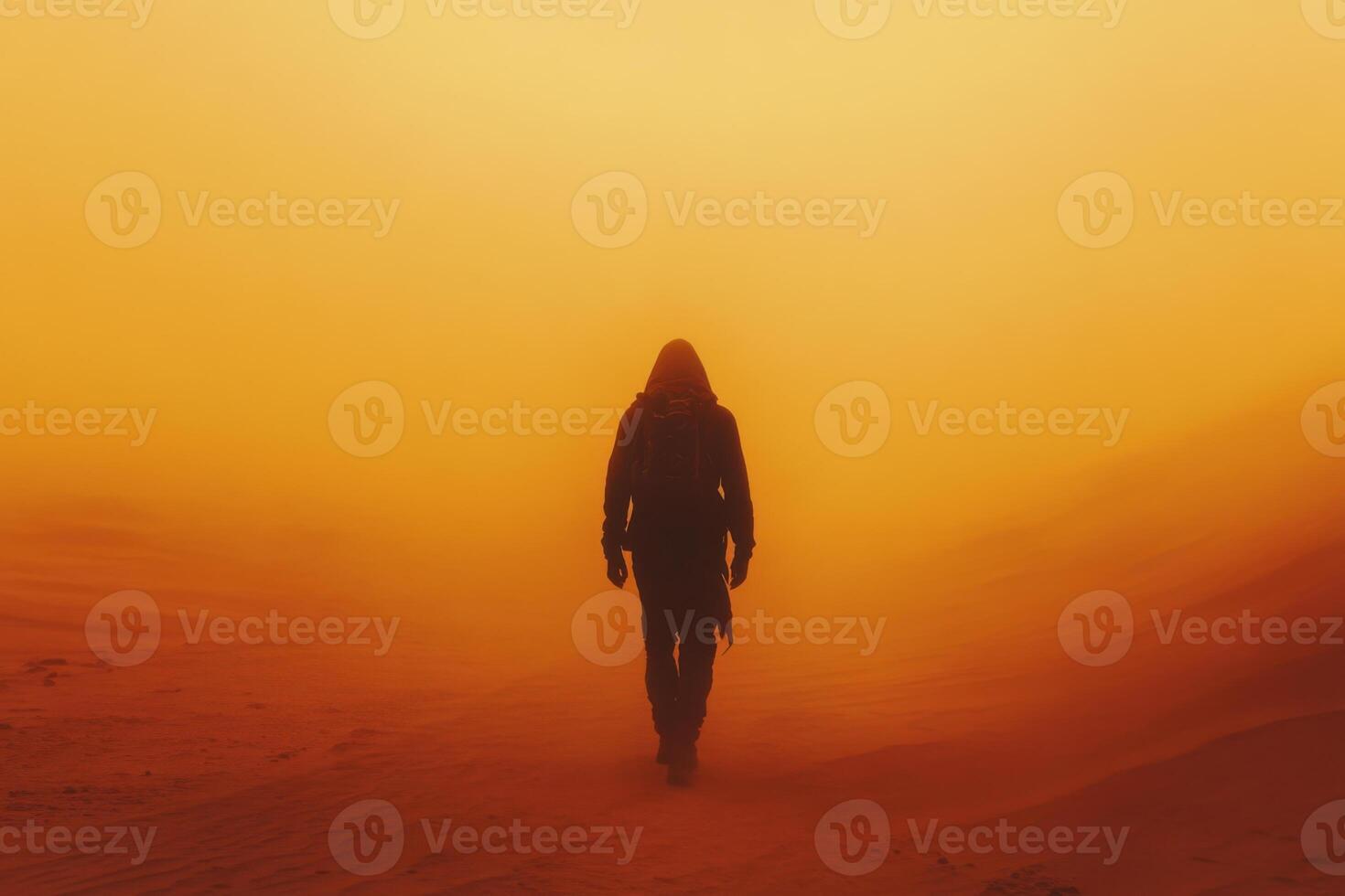 Person with hood walks alone in desert sand. Orange fog covers ground during the day. Walking figure with backpack faces forward. photo