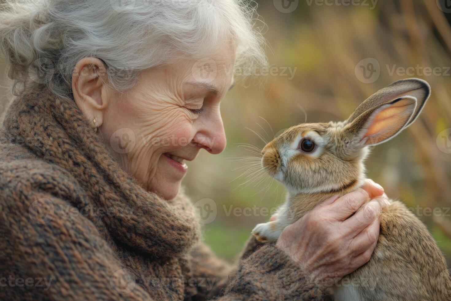 Smiling older woman in warm sweater holds a light brown rabbit. Close up view of animal and person looking at each other outdoors. photo