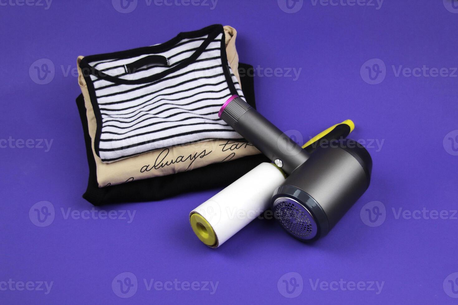 Clothes roller and lint machine are lying on clothes and purple background. In the background are several T-shirts of different colors. photo