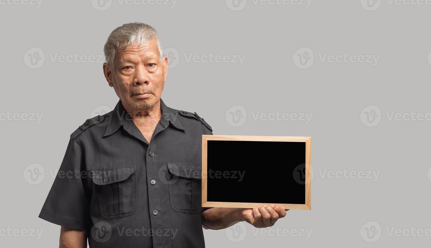 An elderly Asian man holding a mini blackboard and looking at the camera with a smile while standing on a gray background. Space for text. photo