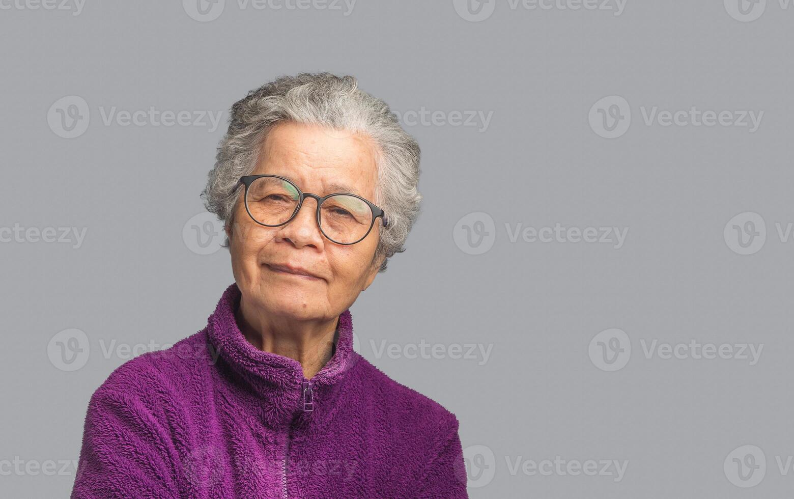 Beautiful elderly Asian woman wearing glasses, looking at the camera with a smile while standing on a gray background. photo