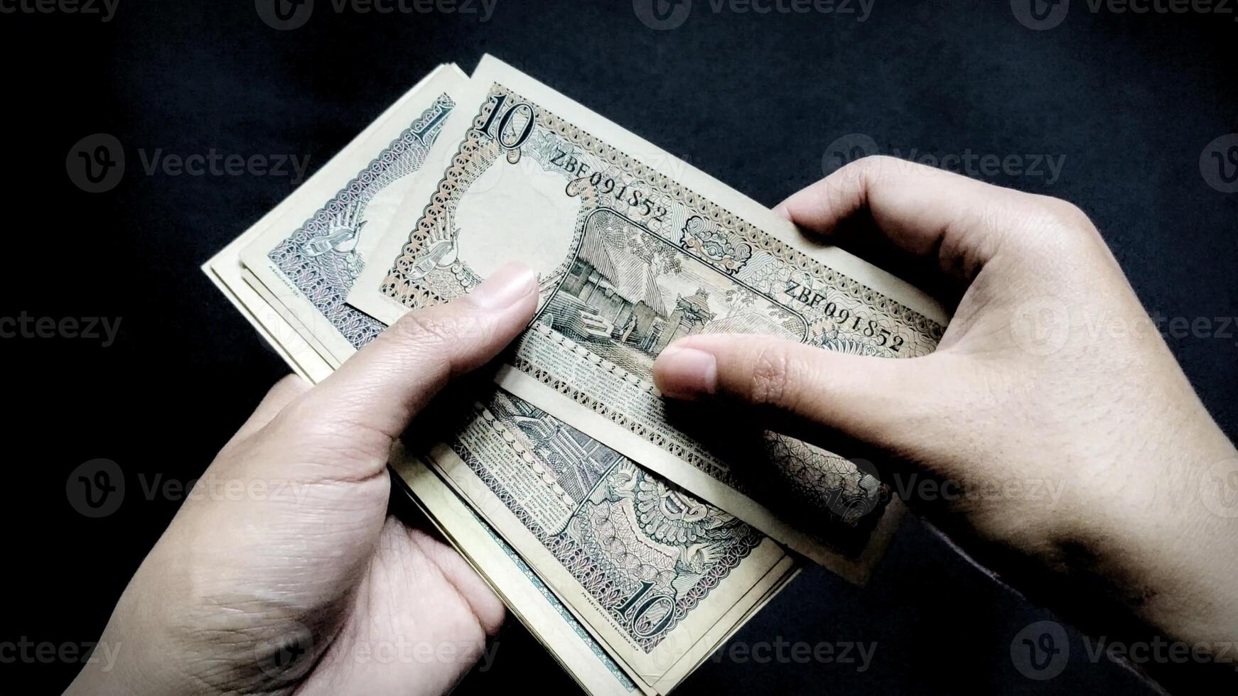 Low light concept of a man holding some old rupiah Rp.10 notes. Isolated on a black background, close up view photo