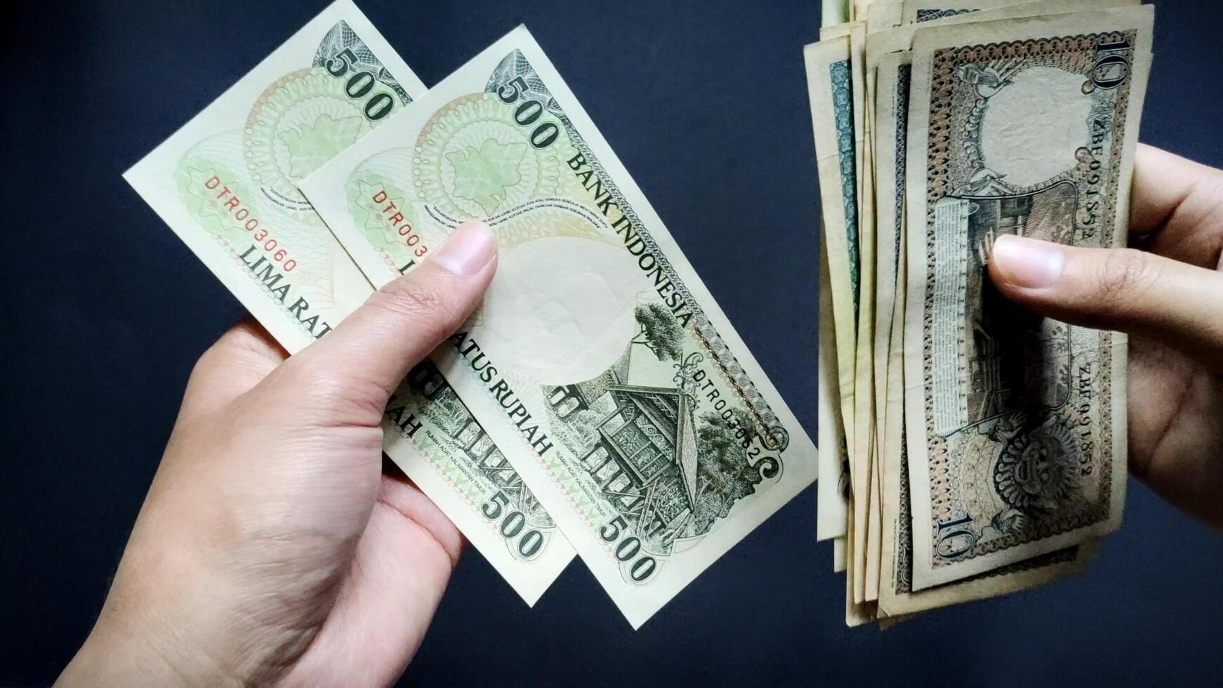 A person's hand is seen counting old rupiah banknotes of Rp10 to Rp500, isolated on a black background. Close-up view photo
