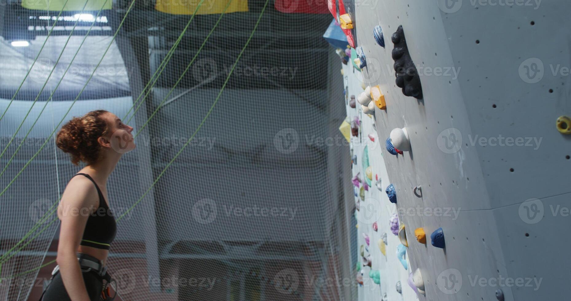 Young woman athlete looks up while standing in front of a climbing wall photo