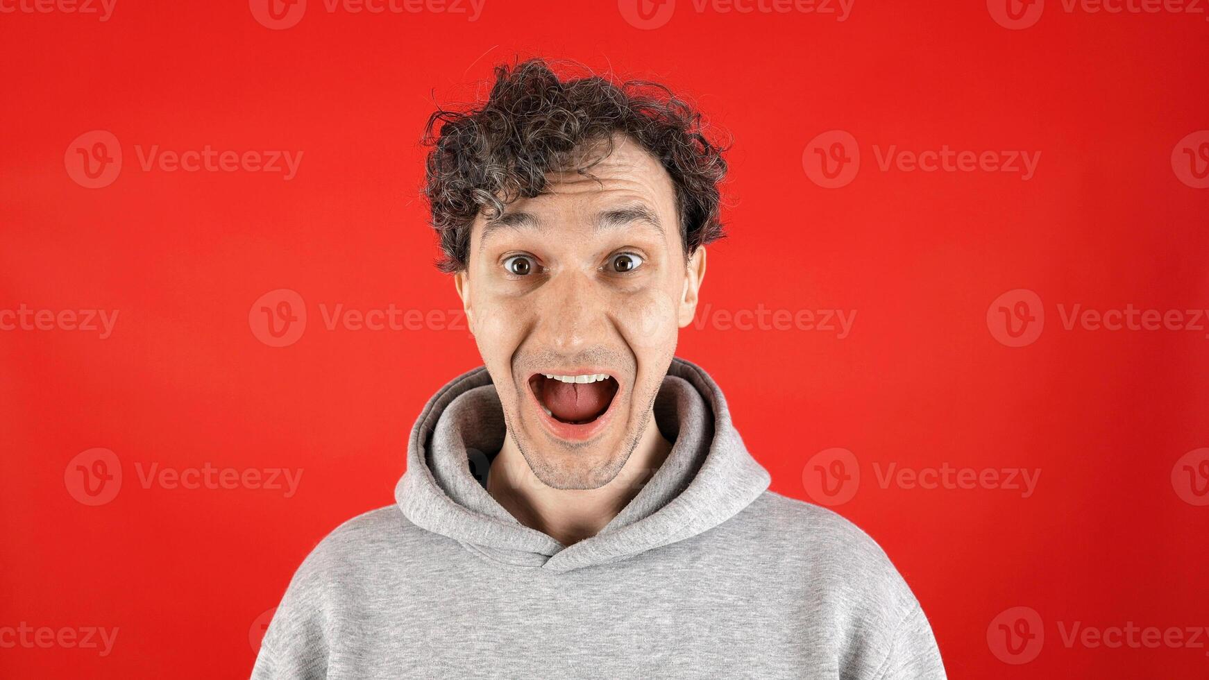 large close up portrait young curly man, joyfully surprised, holding his face with mouth open, pronouncing word wow on studio red background. Concept - discounts, Black Friday, excitement, advertising photo