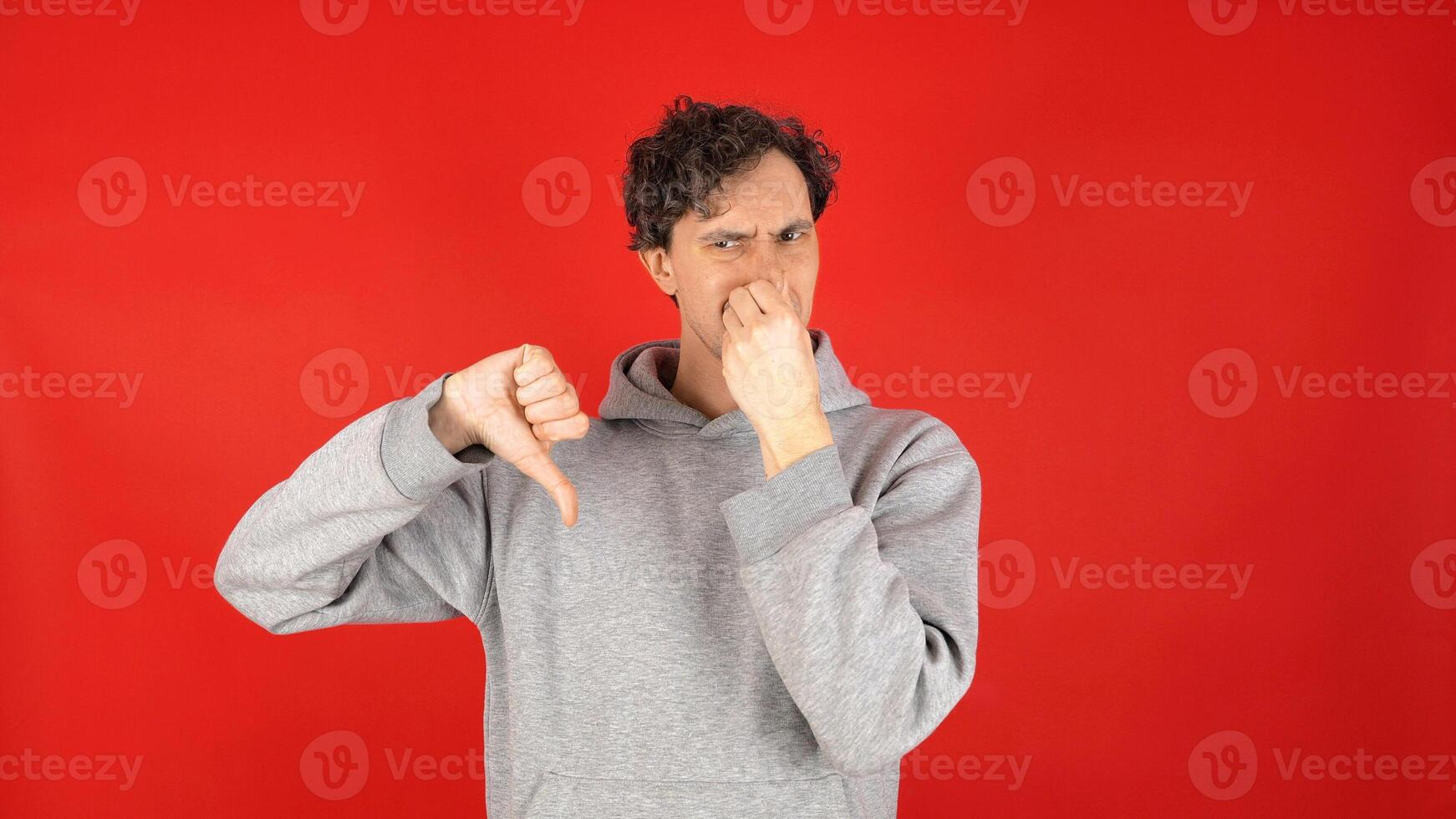 Portrait displeased unhappy young adult curly man squeezing nose fingers, waves hand in front reacting gross, smelly odor. Isolated on red background studio shot. Capturing strong reaction to smell. photo
