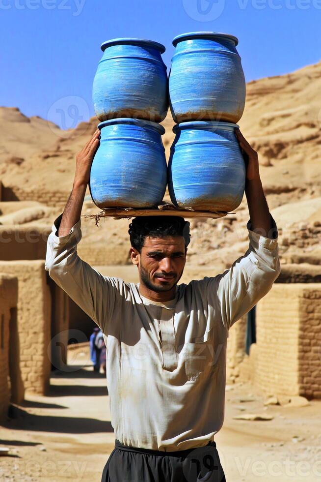 A man carries four blue pots on his head in a desert village setting. photo