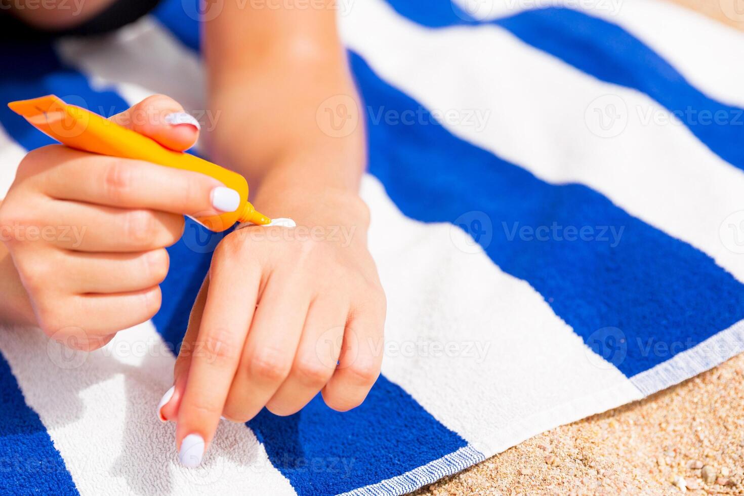 Young woman lying on striped towel on the sand at the beach and applying sun cream on her hand photo