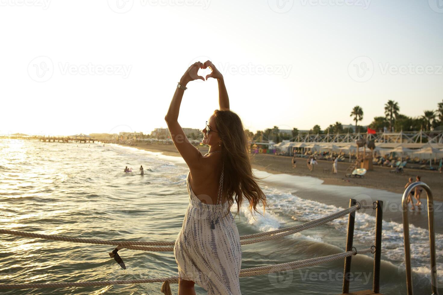 Woman on Pier Making Heart Gesture Overlooking Sunset Beach and Ocean Waves photo