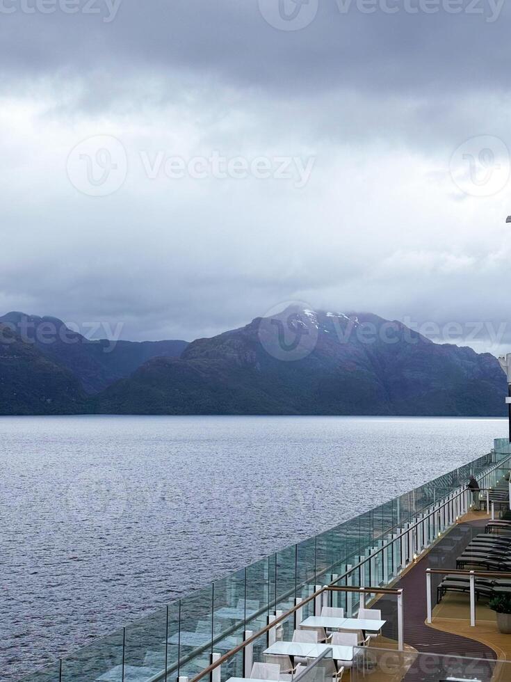 Cruise Ship Deck Overlooks Calm Fjord and Snowy Mountain Under Cloudy Sky photo