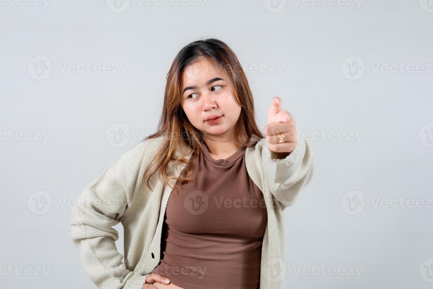 A young woman points directly at the camera with a confident and engaging expression, wearing a cozy sweater. She connects with the viewer against a neutral background photo