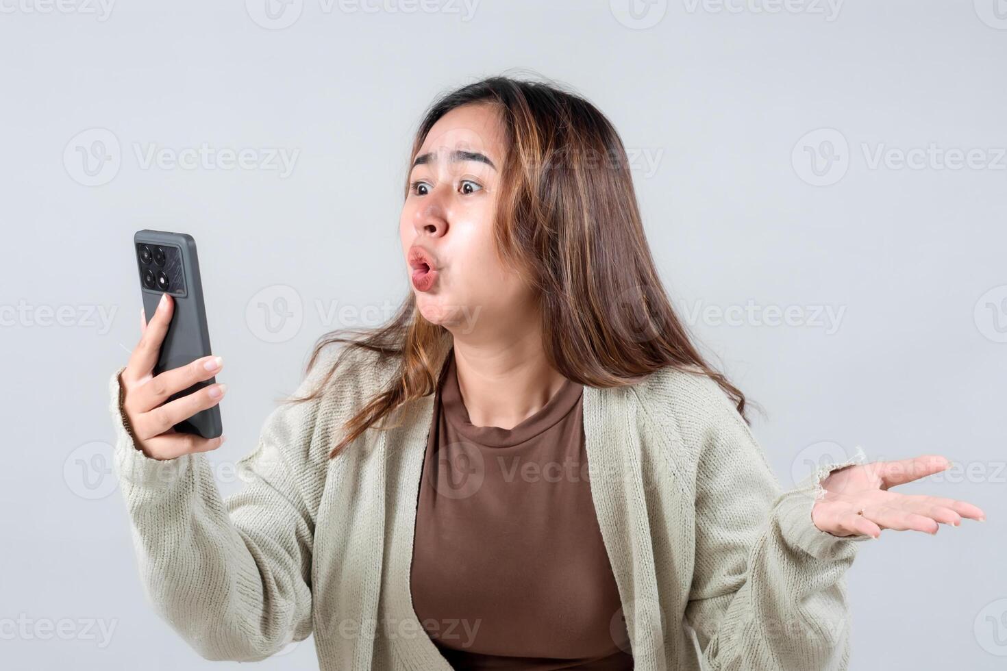 young woman expresses frustration while looking at her smartphone, showcasing a strong emotional reaction in a neutral background. photo