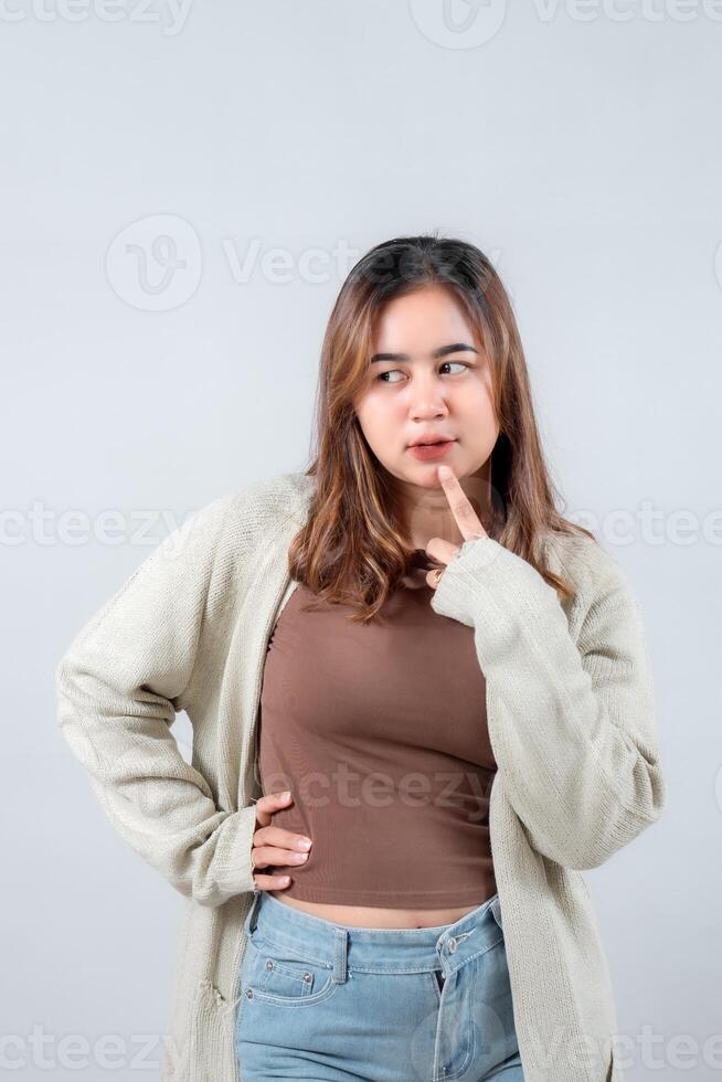 young woman in a cozy sweater, with a thoughtful expression, touches her chin while looking to the side against a neutral background photo