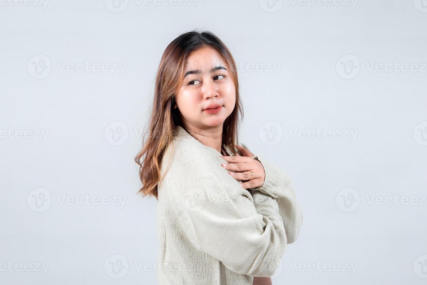 smiling young woman with a warm expression, wearing a cozy sweater holds her hands close to her heart, radiating kindness and positivity against a neutral backdrop. photo