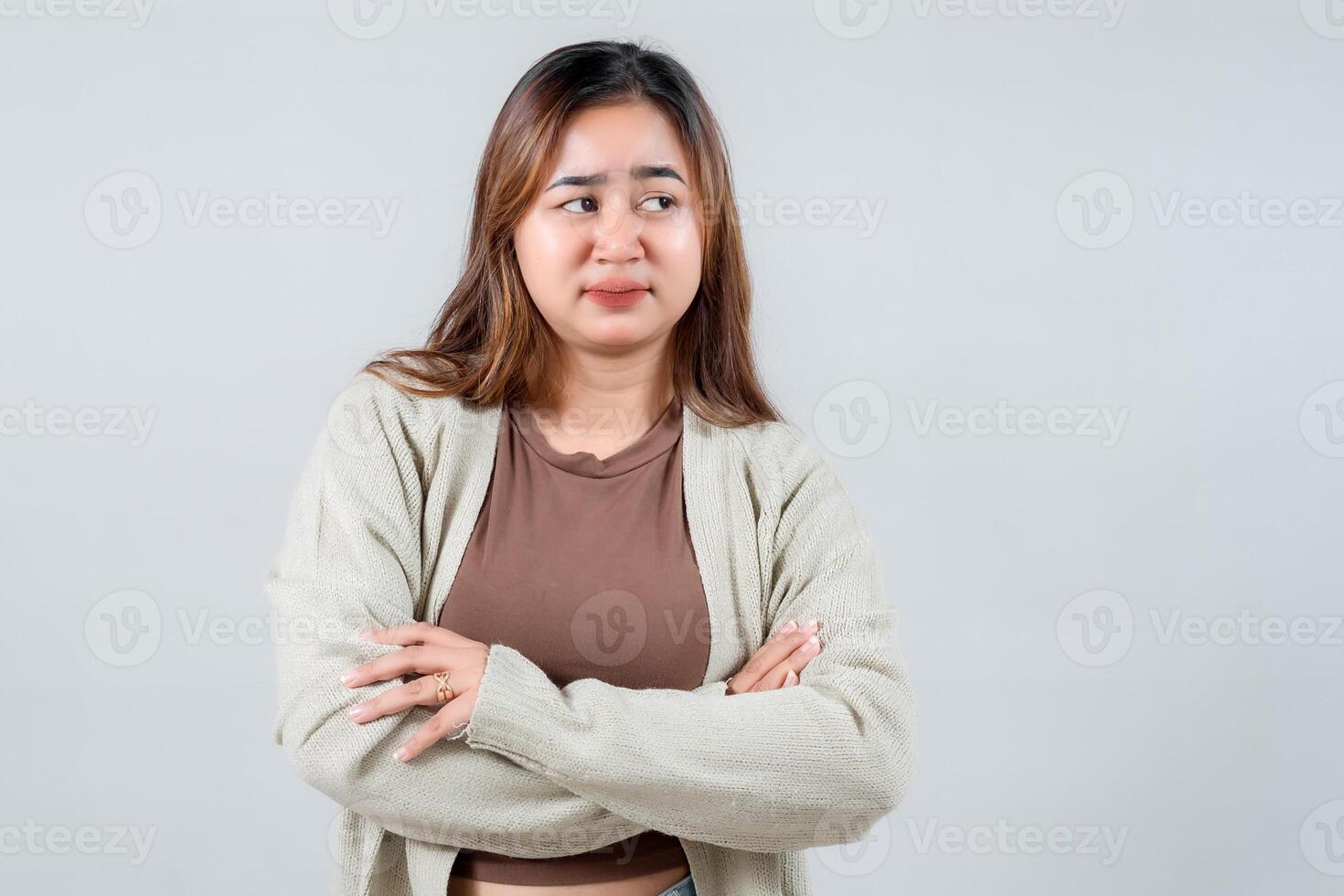 A young woman stands with her arms crossed, with a serious and slightly skeptical expression on her face as she looks to the side. Conveying a sense of contemplation or mild disagreement photo