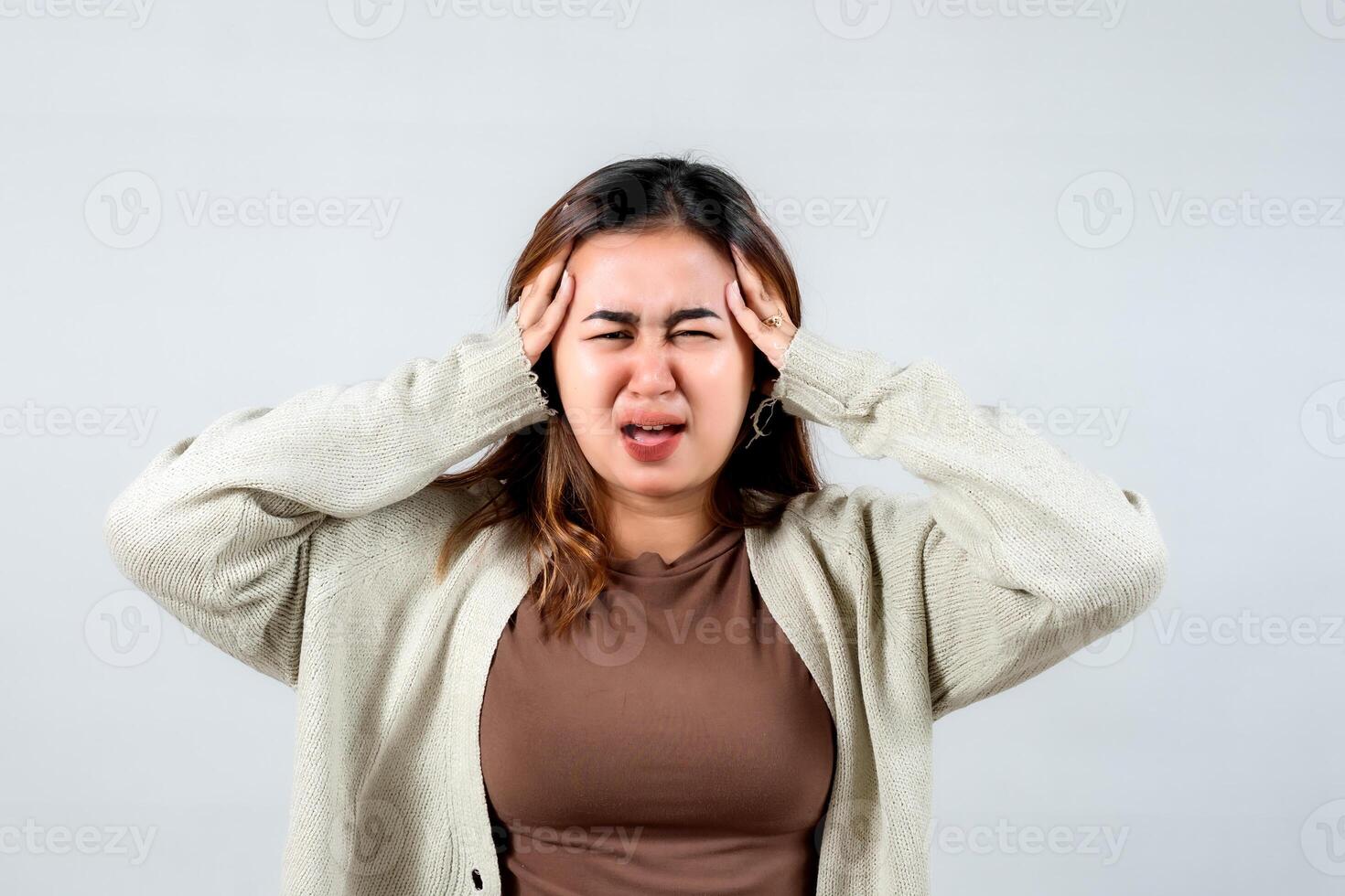 young woman holds her head in her hands, her face expressing pain and distress. Wearing a cozy sweater, she conveys a strong sense of headache or emotional stress against a simple background. photo