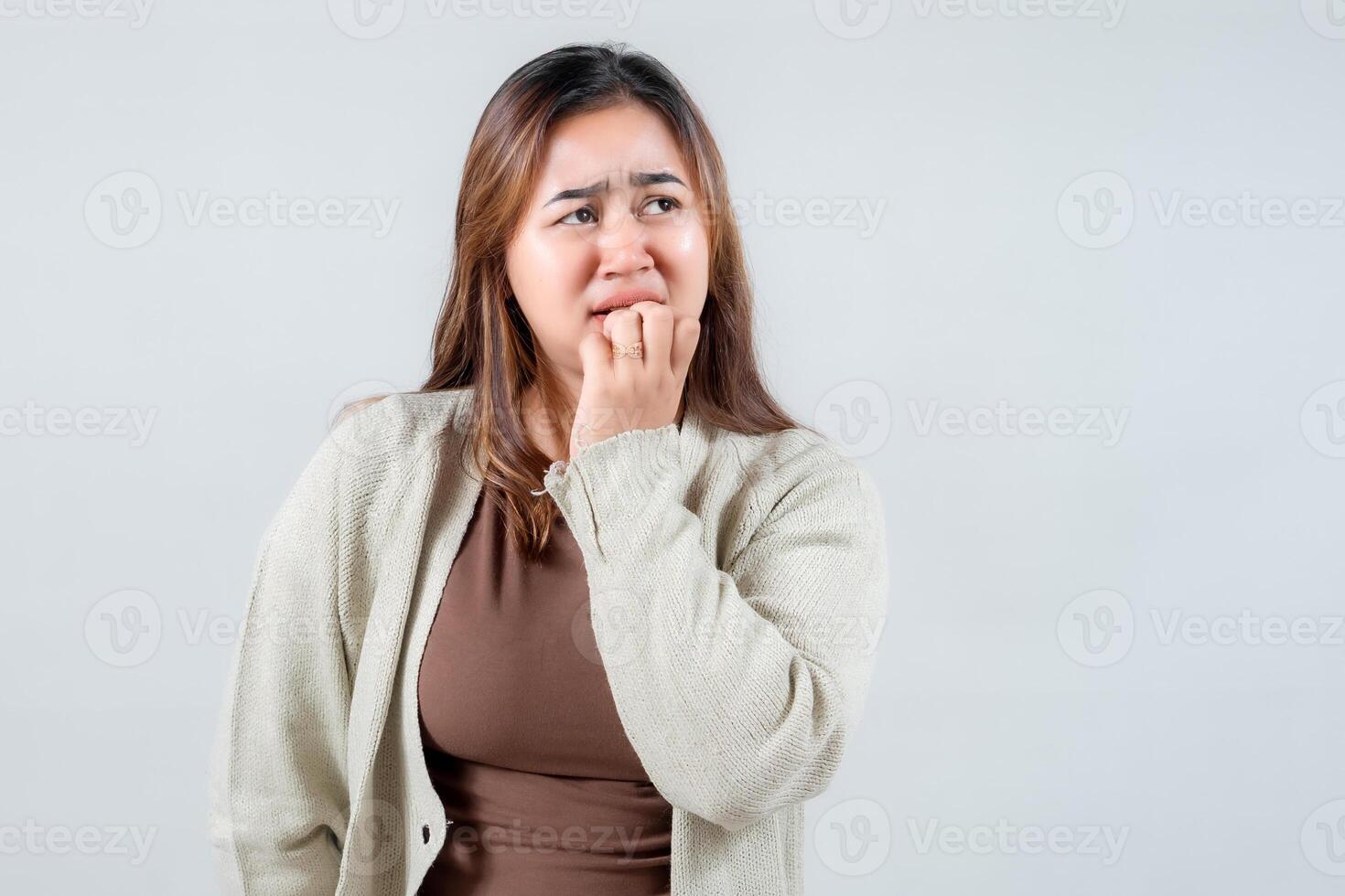 A young woman with long hair looks concerned while biting her nails, wearing a cozy sweater. Her expression reflects anxiety and contemplation against a simple gray, capturing a relatable moment photo