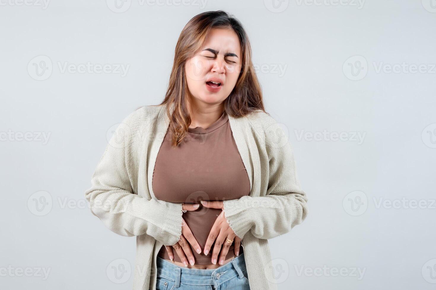 woman grimaces uncomfortably while holding her stomach, wearing a comfortable sweater. Her expression suggests pain or anxiety, capturing a moment of incomprehensible distress. photo
