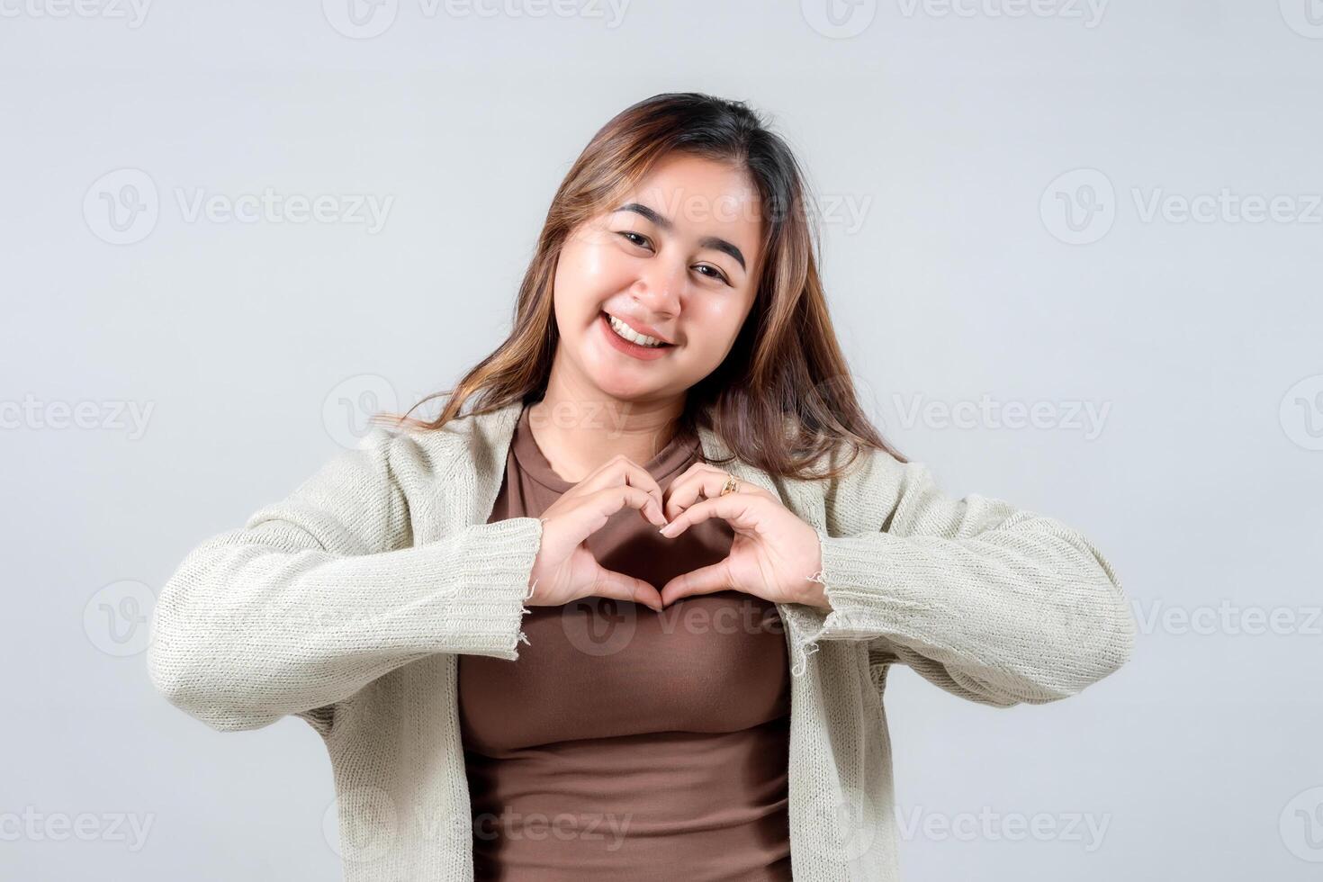 A smiling young woman in a cozy sweater makes a heart shape with her hands, expressing love, affection, and kindness against a neutral background. photo
