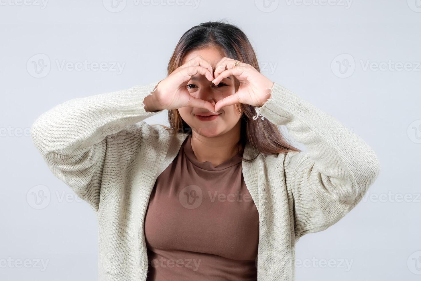 A young woman in a cozy sweater forms a heart shape with her hands, expressing love, care, and affection against a neutral background. photo