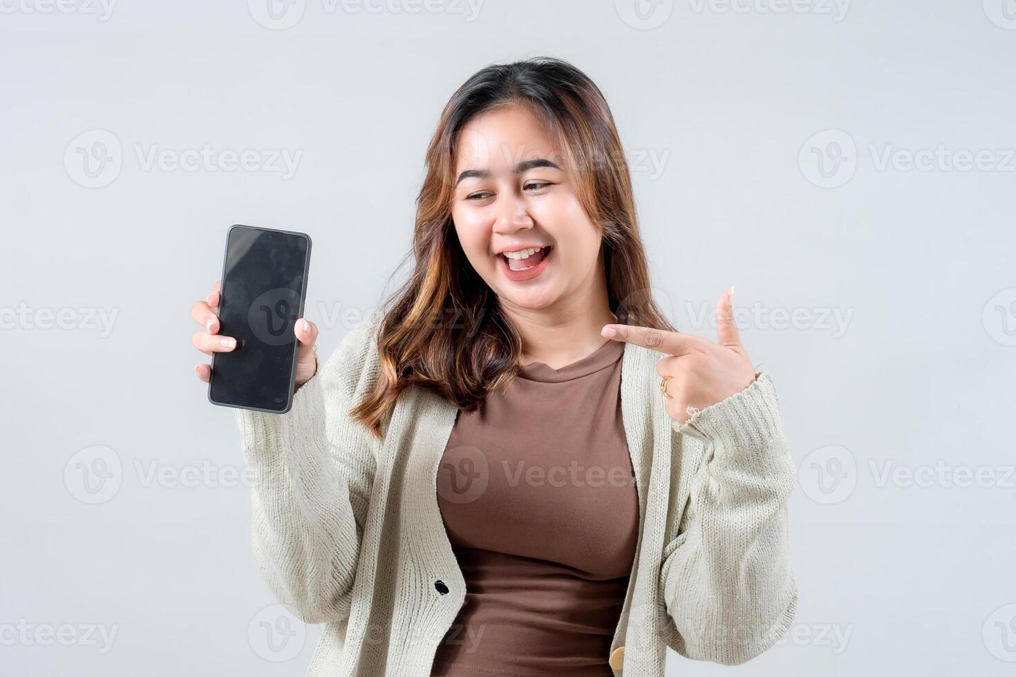A young woman smiles and points at her smartphone, showcasing excitement and enthusiasm while holding the device in a neutral background. photo