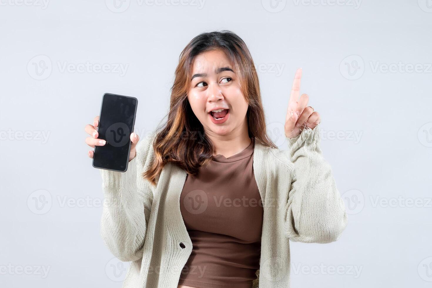 A young woman holds a smartphone and points upwards, suggesting an idea or promotion with a cheerful expression against a neutral background photo